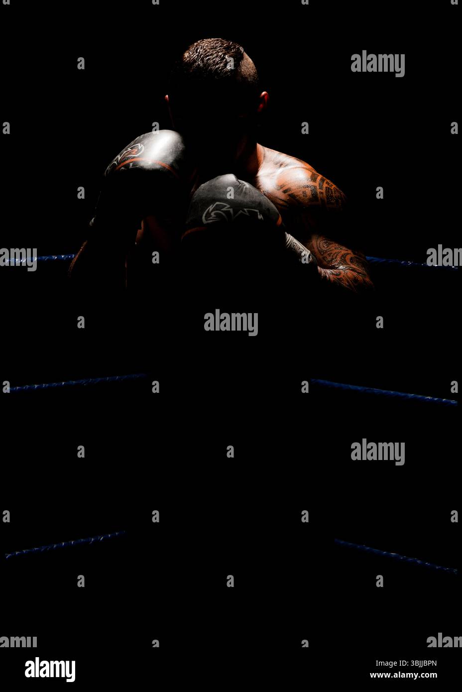 Ein männlicher Boxer mit Tätowierungen, sein Gesicht verdeckt von Schatten, steht in einem schwach beleuchteten Boxring in einer defensiven Haltung. Der Fokus liegt auf seinem Oberkörper Stockfoto