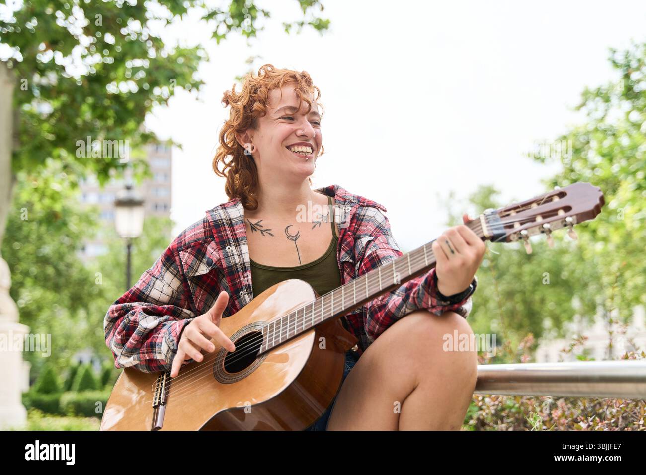 Glückliche junge Frau mit lockigen roten Haaren und Tattoos, die ihre Akustikgitarre in einem Stadtpark spielt und ihr musikalisches Talent und ihre Leidenschaft zum Ausdruck bringt Stockfoto