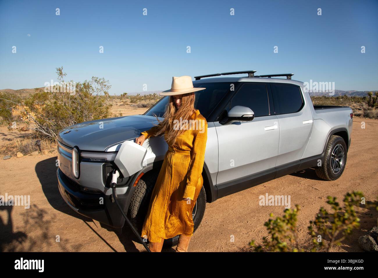 Stilvoll gekleidete Frau, die sich gegen das Laden eines Elektrofahrzeugs lehnt Stockfoto