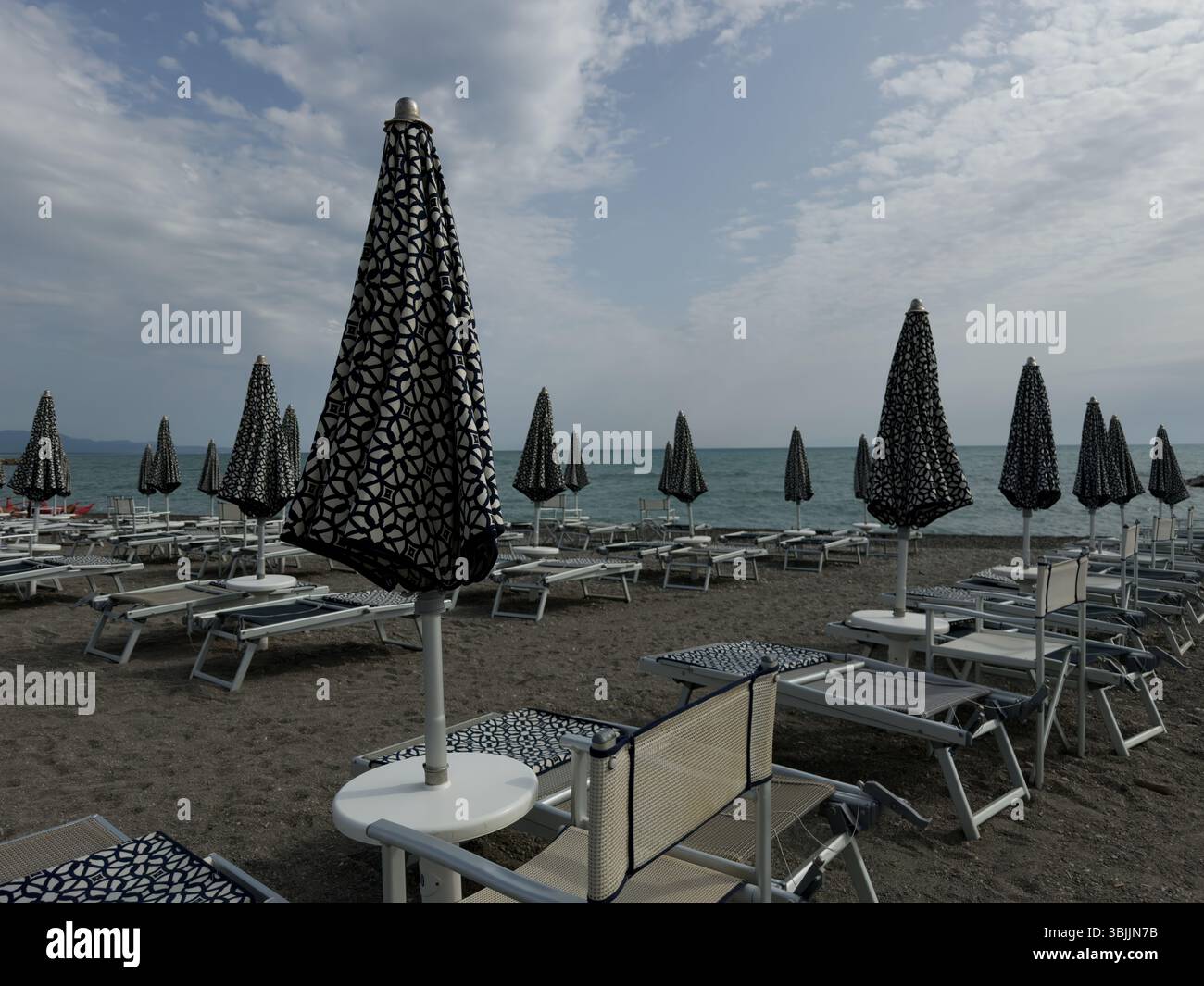 Geschlossene Sonnenschirme und Liegestühle am Morgenstrand vor dem Hintergrund der Küste - Smartphone-aufgenommenes Stockfoto