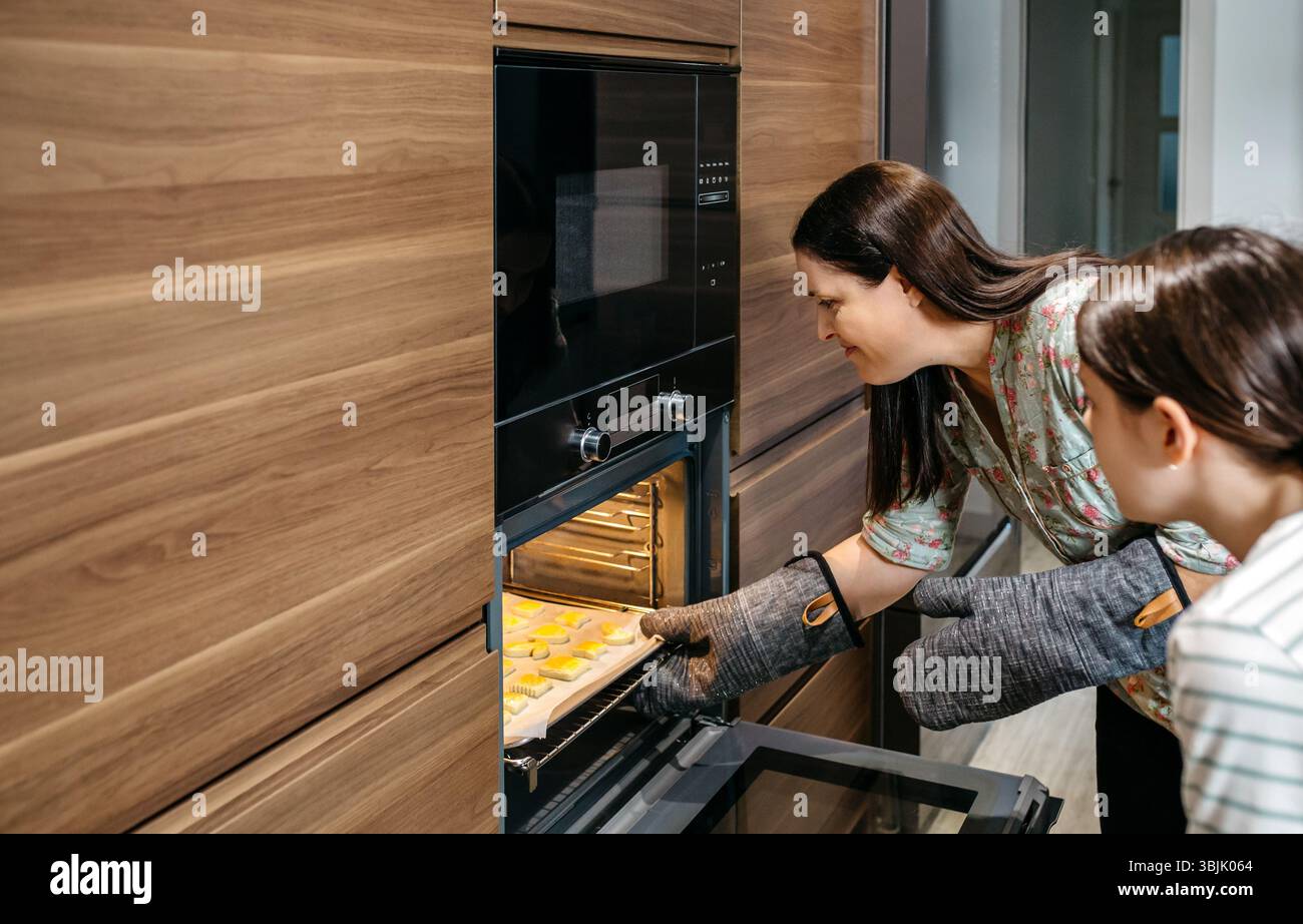 Mom und ihre Tochter nehmen eine Pfanne voller goldener Kekse aus dem Ofen in ihrer Küche, tragen Ofenhandschuhe und arbeiten zusammen, um sich zu kümmern Stockfoto