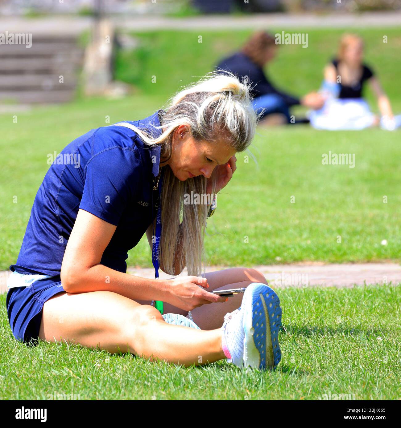 Sportlich gekleidete Frau studiert ihr Telefon im öffentlichen Park, Somerset, England, Großbritannien. Juni 2025. Sommer Stockfoto