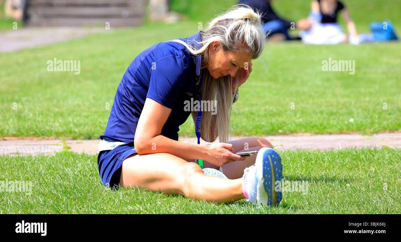 Sportlich gekleidete Frau studiert ihr Telefon im öffentlichen Park, Somerset, England, Großbritannien. Juni 2025. Sommer Stockfoto