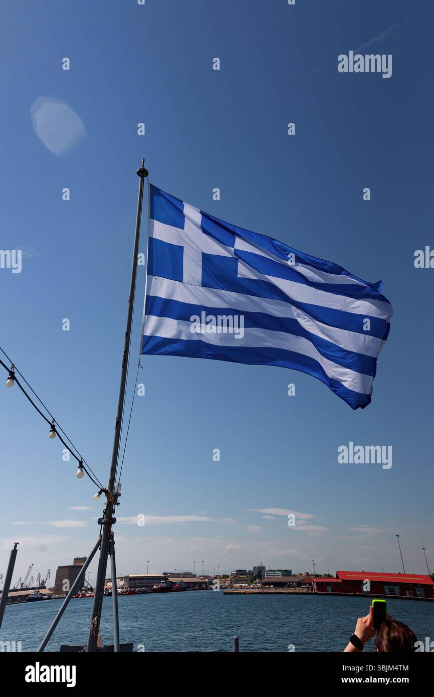 Große griechische Flagge winkt mitten in der Luft über dem Meer mit blauem Himmel während des Tages. Stockfoto
