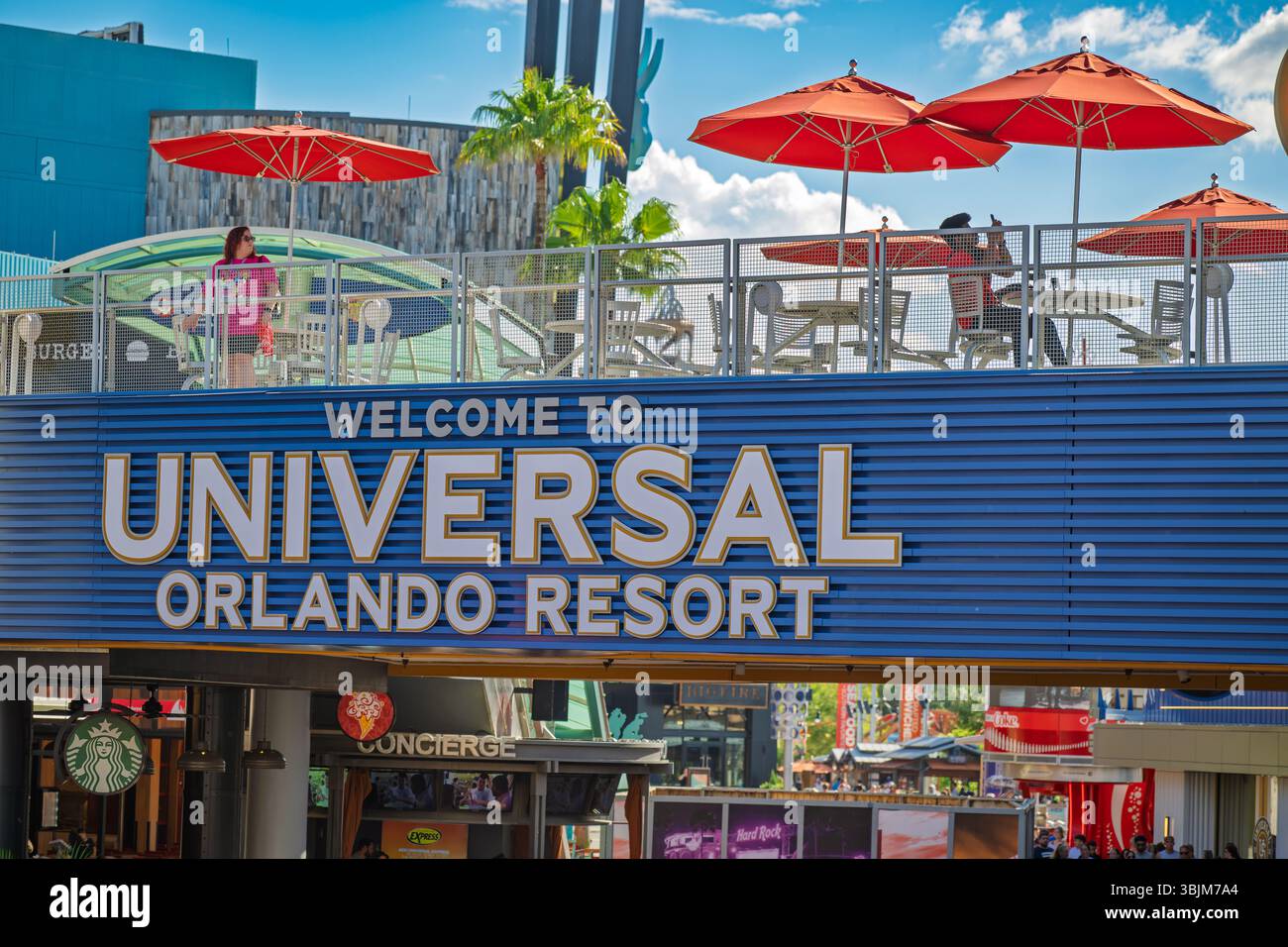 Orlando, Florida, Usa, 13. Mai 2025. Ein Mann sitzt über dem Universal Orlando Resort Schild, unter einem orangefarbenen Schirm, vertieft in sein Handy, während ein Arbeitsauftrag Stockfoto