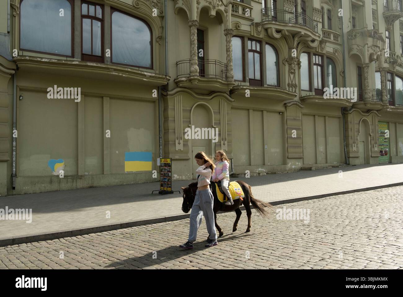 Auf der Deribasovskaja Straße reiten die Menschen vor dem Velyka Moskovska Hotel (derzeit geschlossen). Bekannt für seine neoklassizistische und barocke Architektur, wurde es 2023 zum UNESCO-Weltkulturerbe erklärt. Das historische Zentrum ist weiterhin anfällig für russische Angriffe. Stockfoto