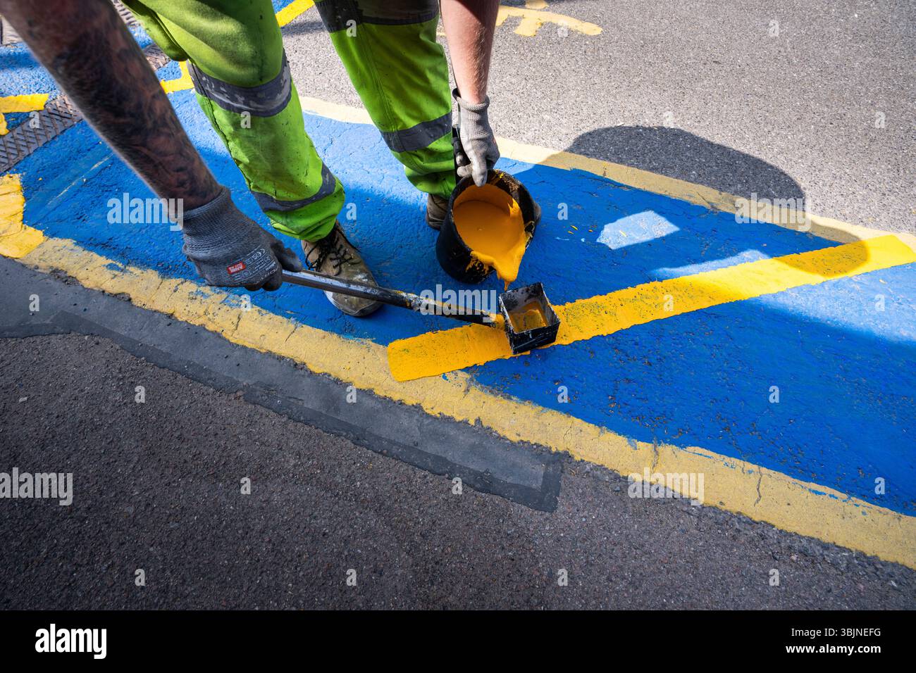 Der Arbeiter gießt heiße gelbe Straßenmarkierungsfarbe in die quadratische Führung, um gerade gelbe Linien auf blauem Hintergrund zu machen Stockfoto