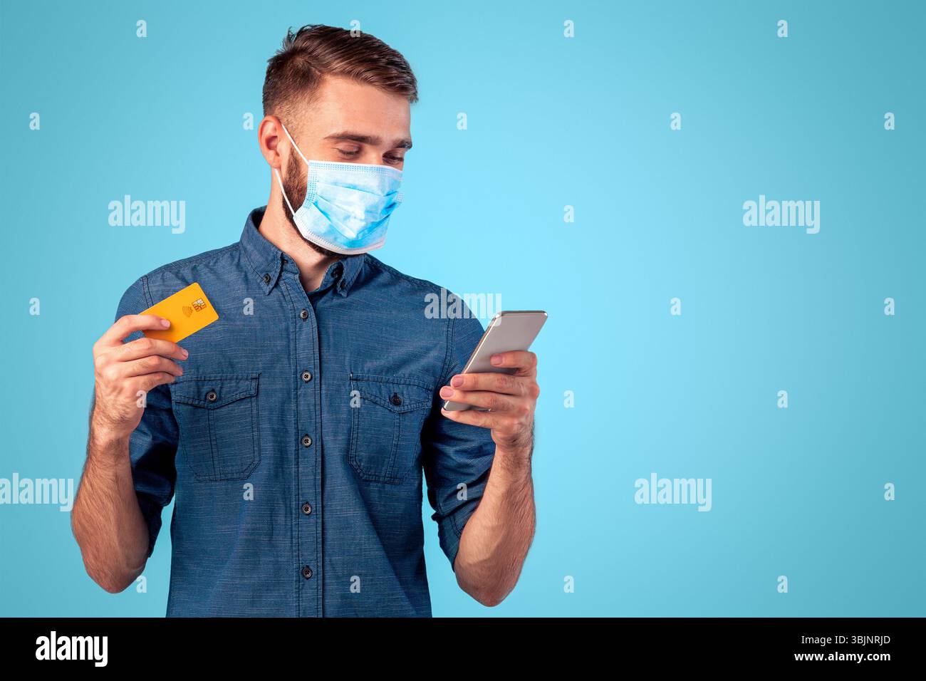 Kontaktloses Einkaufen während der Covid-Isolation. Porträt von Millennial Kerl in Gesichtsmaske hält Smartphone und Kreditkarte Stockfoto