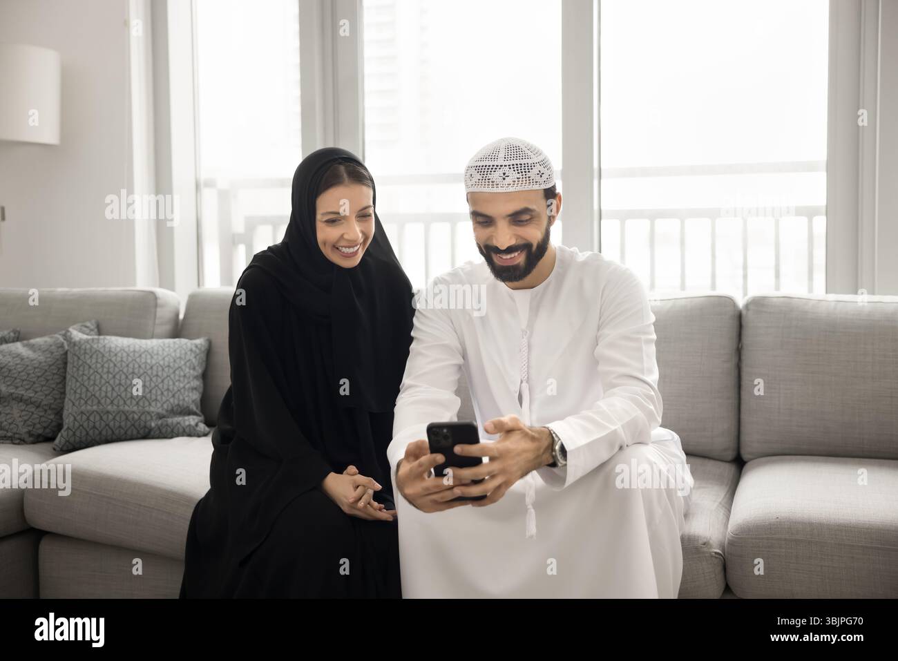 Glückliches arabisches Paar in muslimischer Kleidung mit Smartphone auf dem Sofa Stockfoto