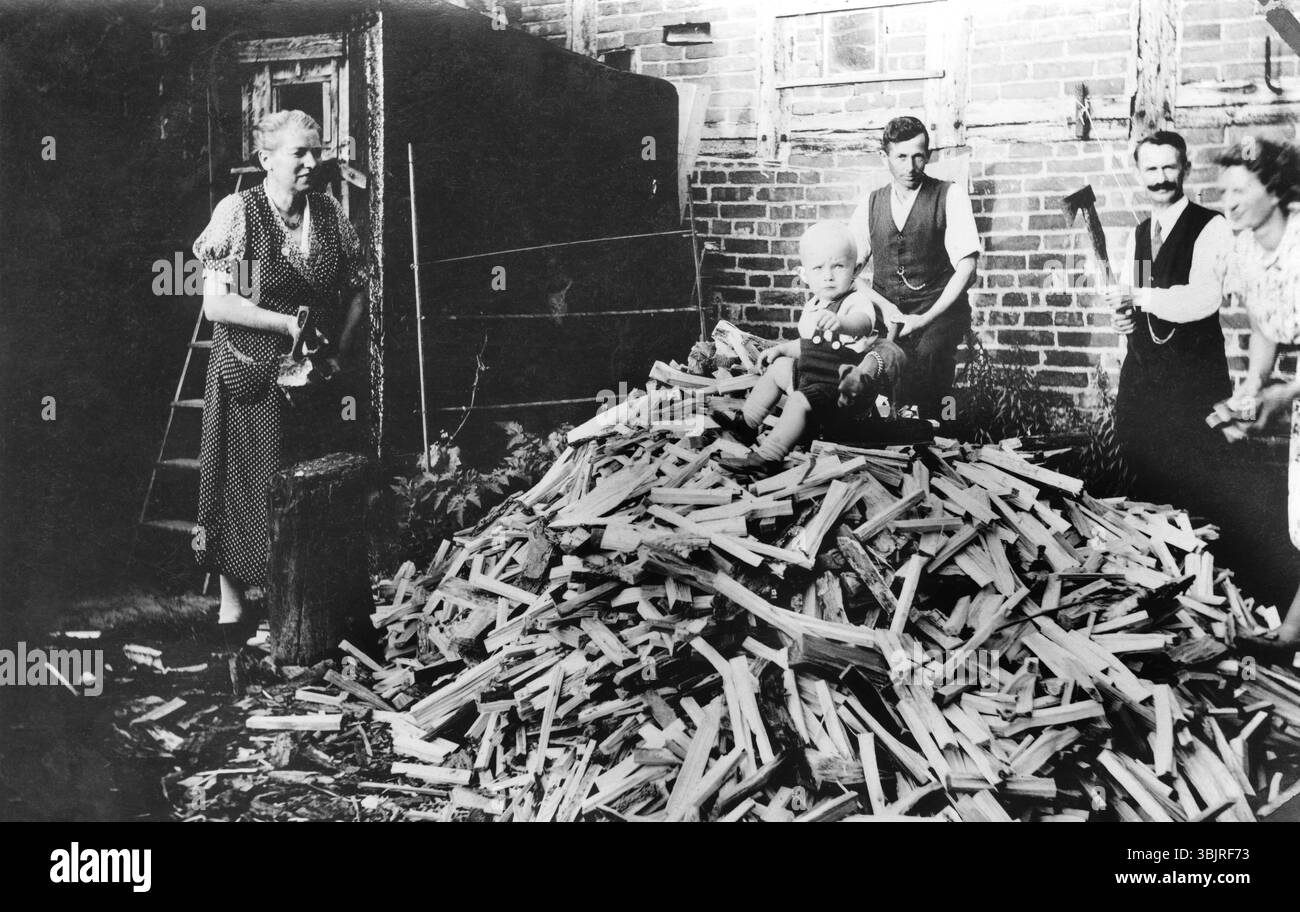 Baby, das auf einem Berg aus Brennholz sitzt, 1930er Jahre, Familie arbeitet zusammen im Hof und hackt Holz, während ein Kind auf dem Holz sitzt, historisches Foto Stockfoto