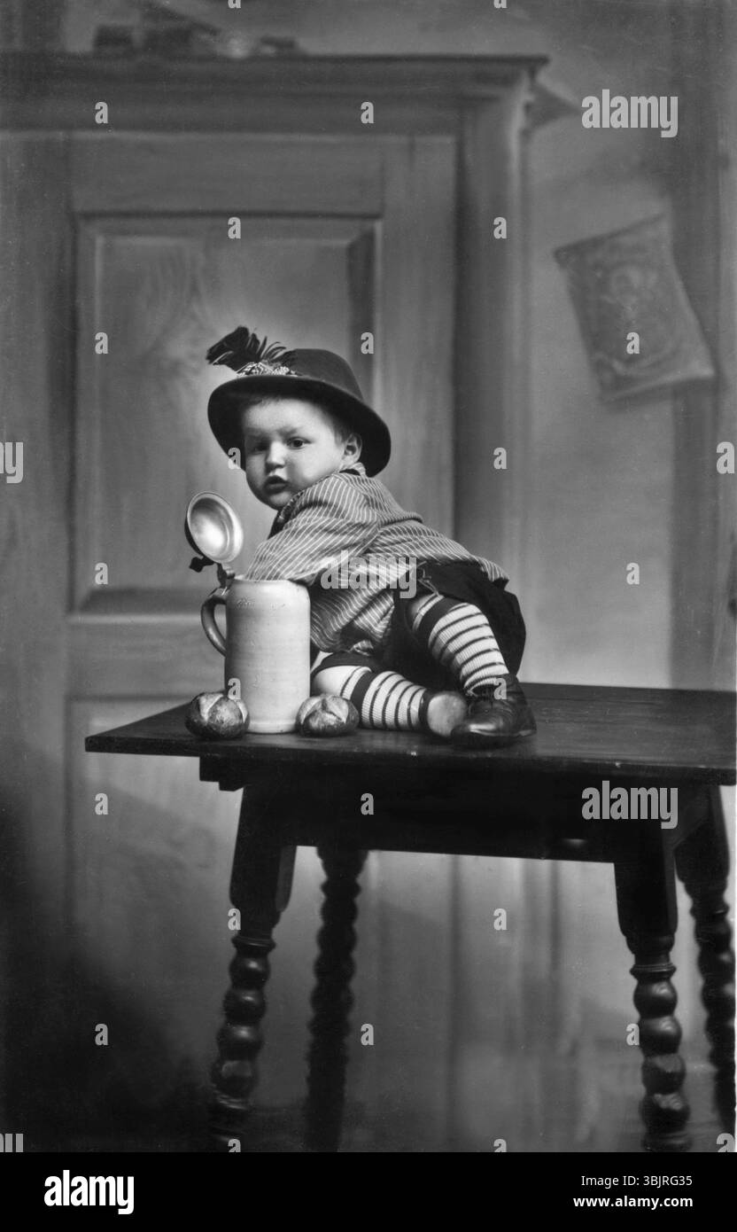 Baby traditionelle Tracht Bierkrug, 1930er Jahre, Kind in traditioneller Kleidung sitzt auf einem Tisch neben einem großen Becher, historisches Foto Stockfoto