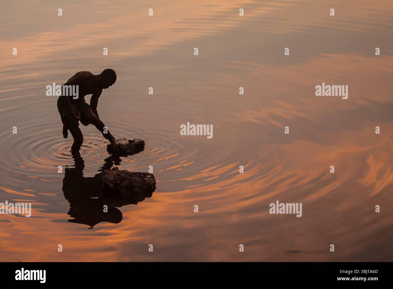Silhouette eines Mannes, der sich die Füße im Niger beim Sonnenuntergang in Ségou, Mali, wascht. Rosa Himmel reflektiert sich auf dem Wasser und lässt Platz für Textüberlagerungen. Stockfoto