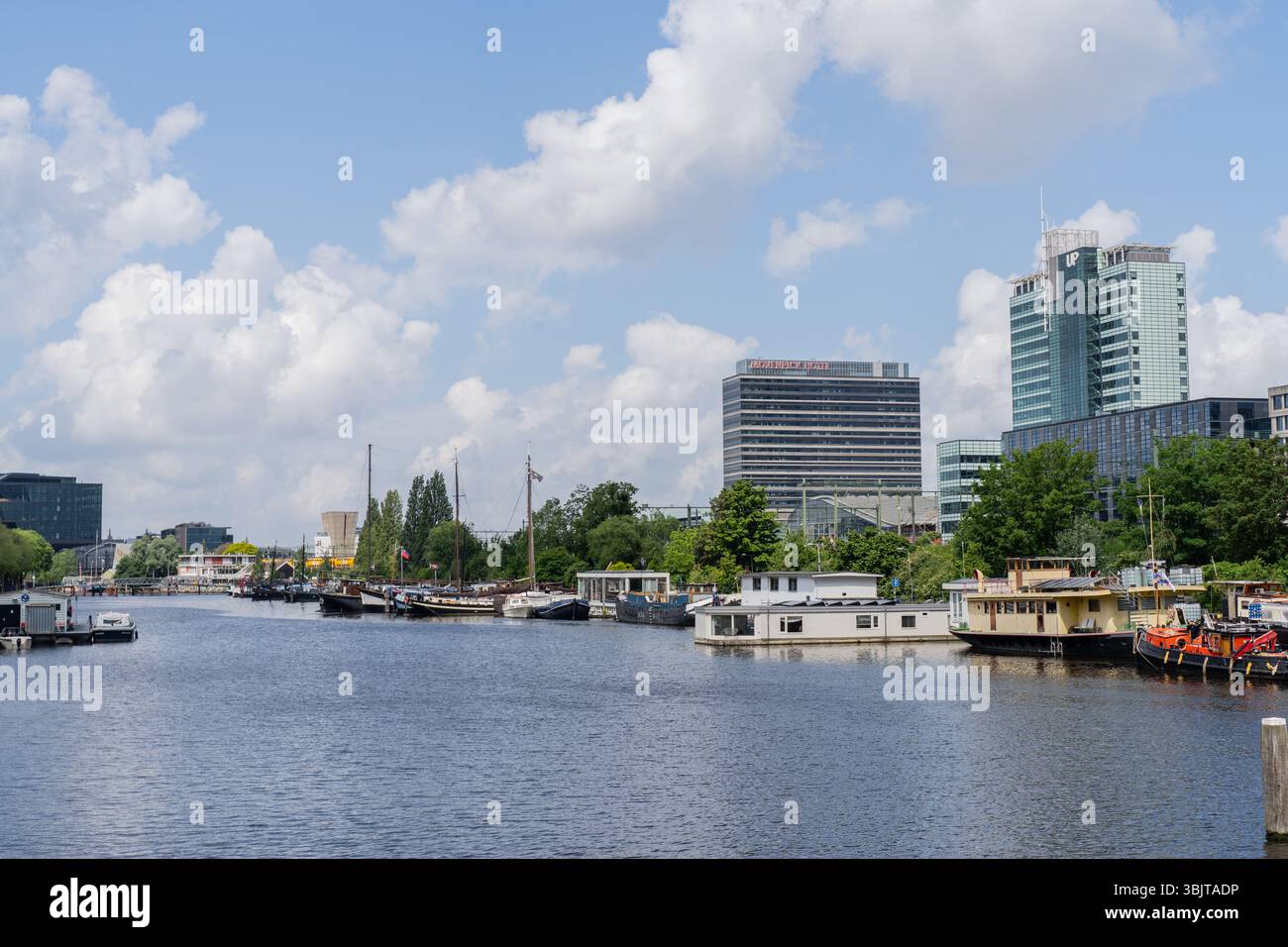 Amsterdam, Niederlande - 7. Juni 2024: Hausboote auf Kanal mit Wolkenkratzern und blauem Himmel Hintergrund in der Hauptstadt Amsterdam. Stockfoto