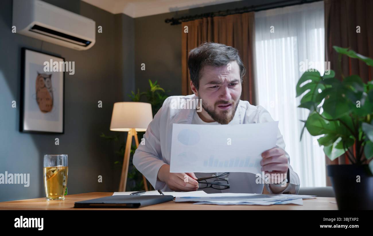 Besorgter Mann in weißem Mantel untersucht gedruckte Diagramme am Schreibtisch zu Hause und weist auf Stress oder Verwirrung bezüglich finanzieller oder medizinischer Daten hin. Stockfoto
