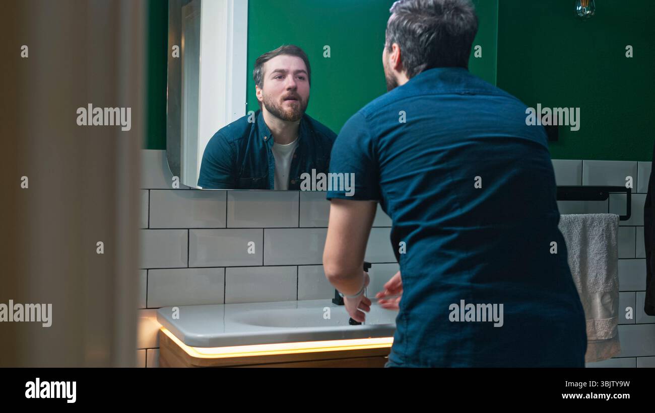 Ein Mann lehnt sich über das Waschbecken und spritzt ihm Wasser auf das Gesicht, während er sich abwäscht. Er sieht konzentriert und auf seine morgendliche Routine gerichtet aus. Er schrubbt h Stockfoto