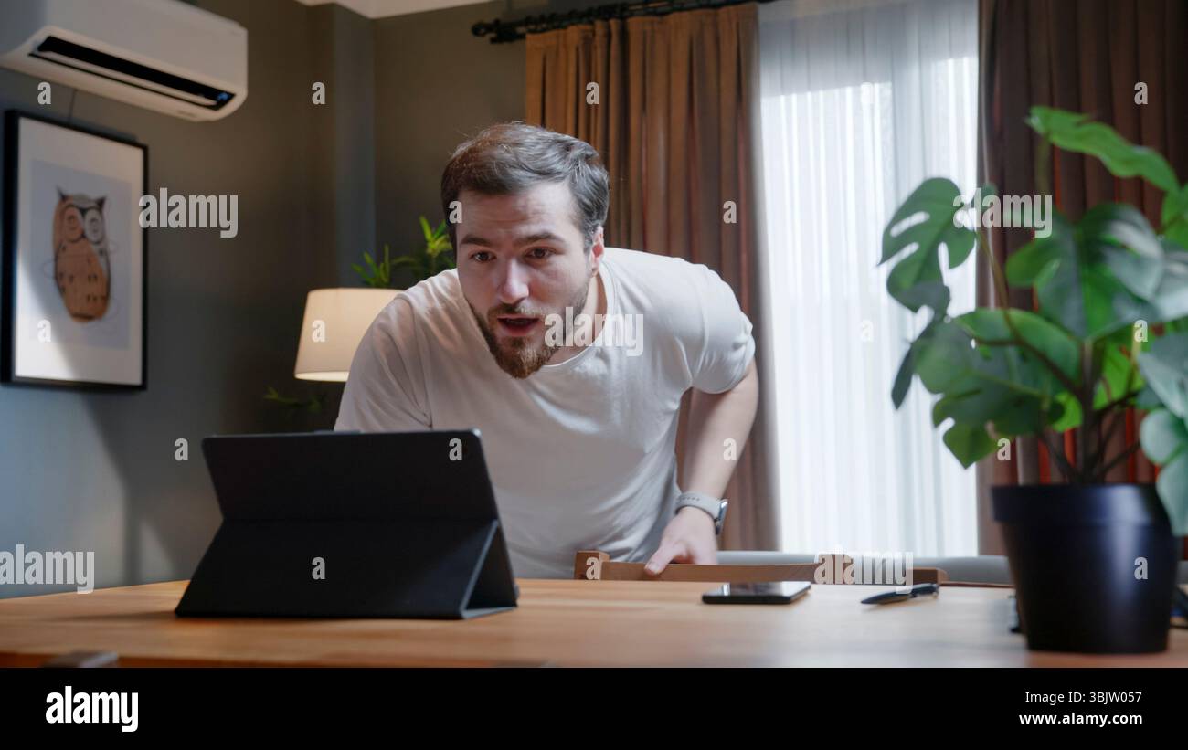 Mann in weißem T-Shirt beugt sich über den Schreibtisch und starrt das Tablet mit einem überraschenden Ausdruck in einem gemütlichen Home Office-Ambiente an. Stockfoto