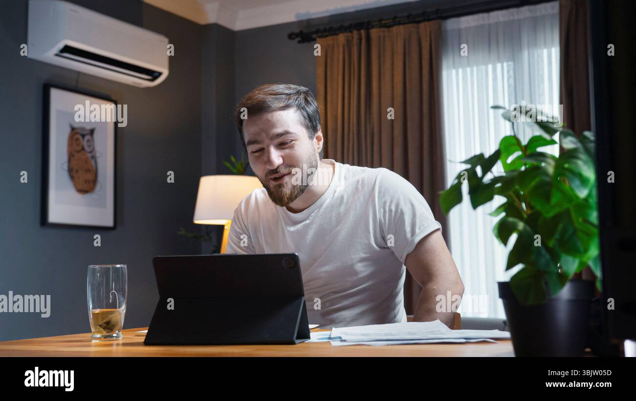 Junger Mann in weißem T-Shirt lächelt und nimmt an einem Videogespräch auf dem Tablet Teil, sitzt mit Papieren und Tee am gemütlichen Schreibtisch. Stockfoto