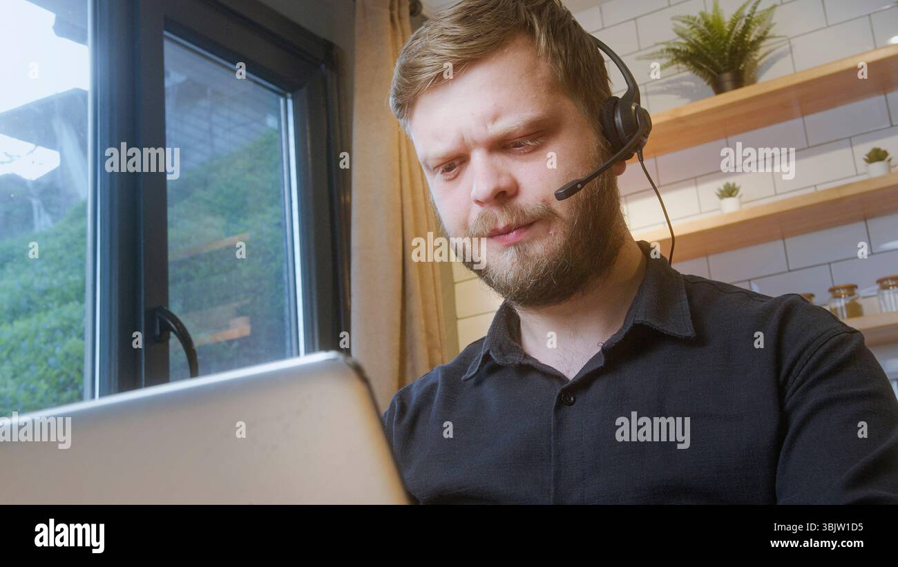 Ein bärtiger Mann, der ein Headset trägt, schaut mit einem verwirrten oder besorgten Ausdruck auf sein Notebook, wahrscheinlich während eines virtuellen Meetings oder eines technischen Supports. Stockfoto