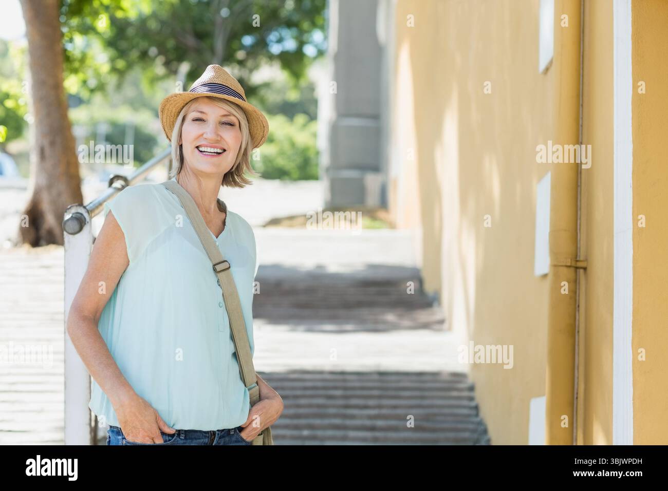 Frau mit Stroh-Fedora-Hut und Canvas-Umhängetasche, die auf sonnendurchfluteten Stufen am gelben Gebäude steht Stockfoto