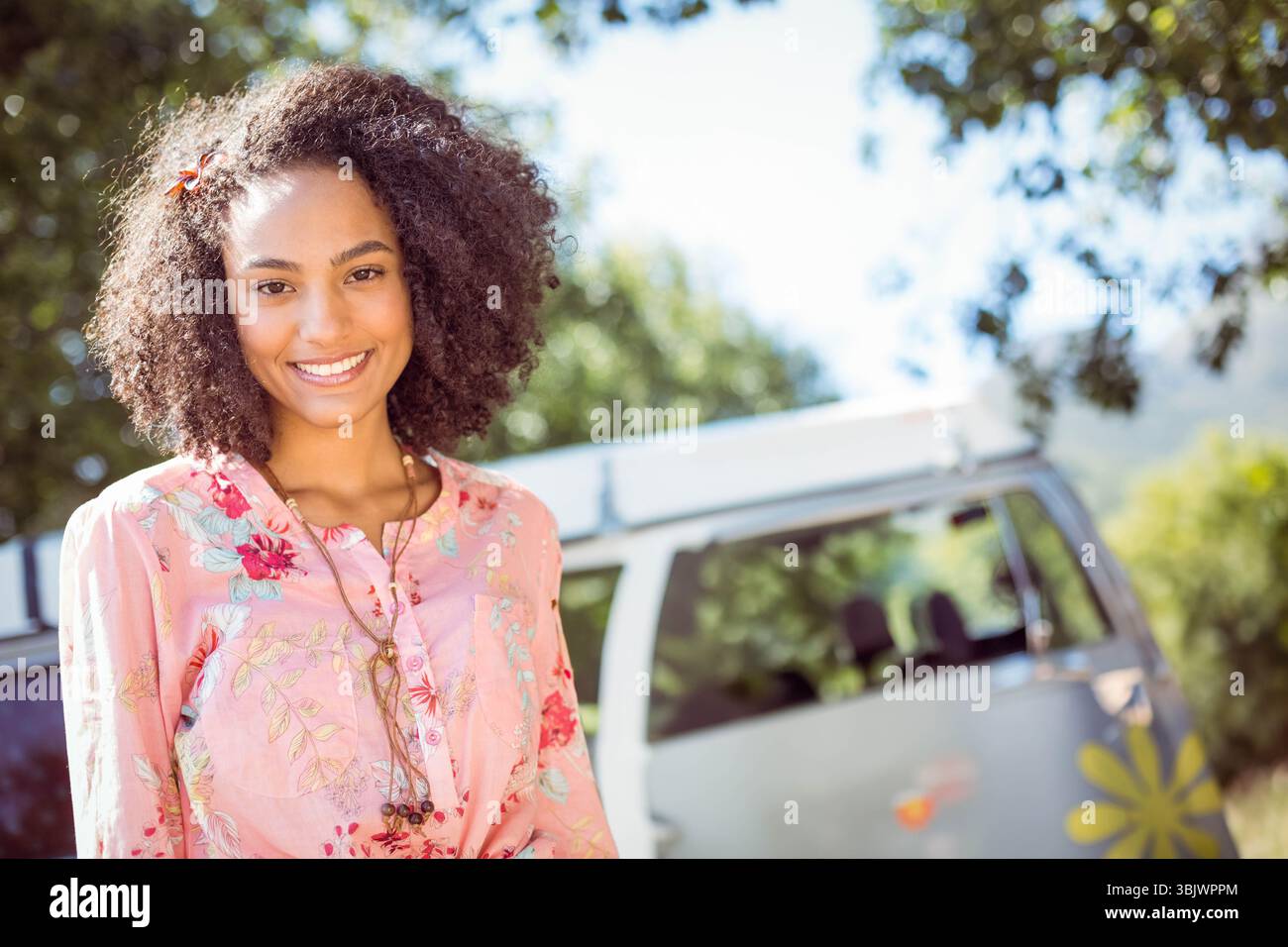 Afroamerikanerin mit rosafarbenem Hemd neben einem Retro-Wohnwagen mit Blumenaufklebern auf dem Campingplatz Stockfoto