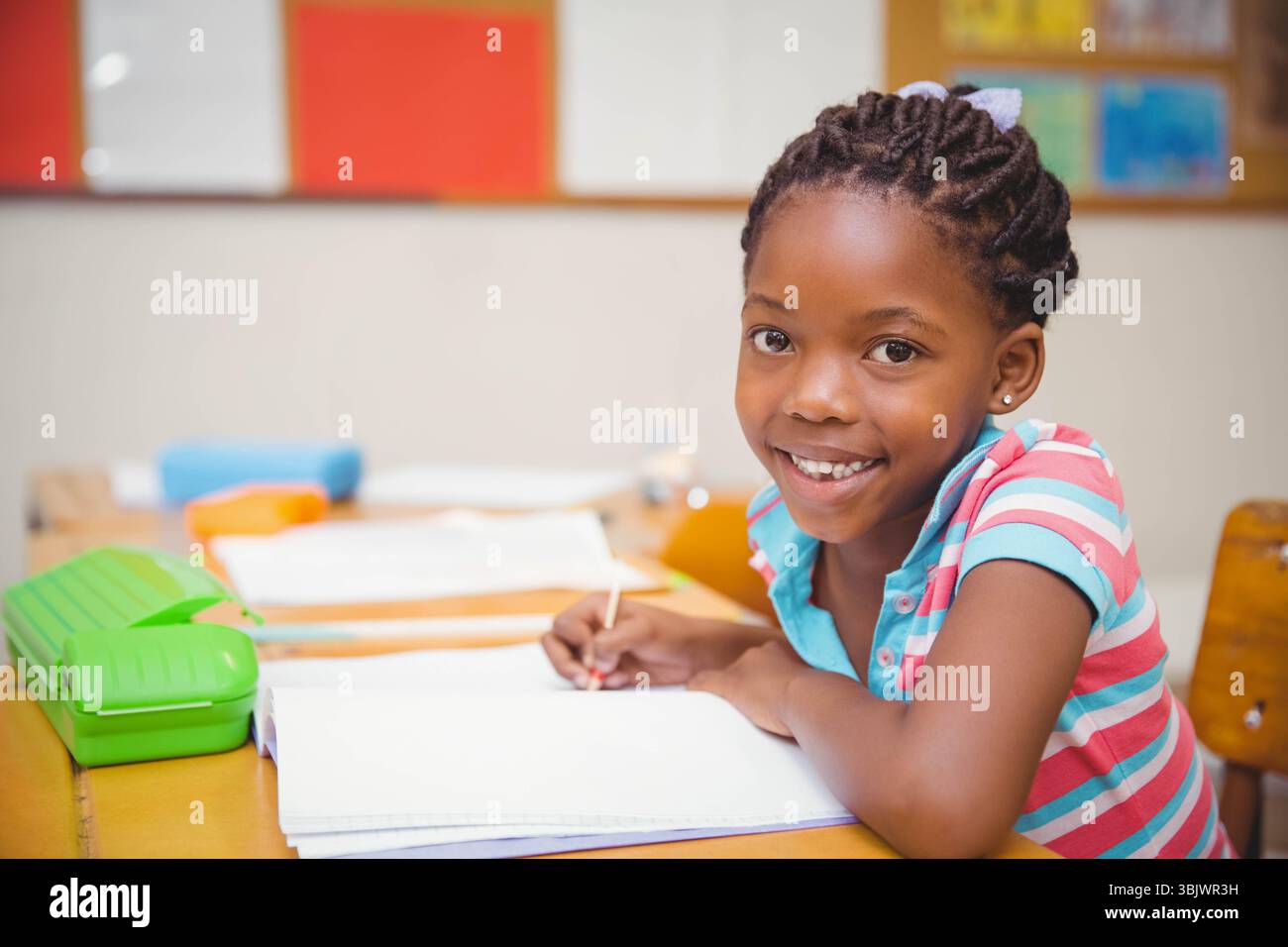 Afroamerikanisches Mädchen sitzt am Schulpult und schreibt in Notizbuch in der Nähe des grünen Bleistiftetui, Kopierraum Stockfoto