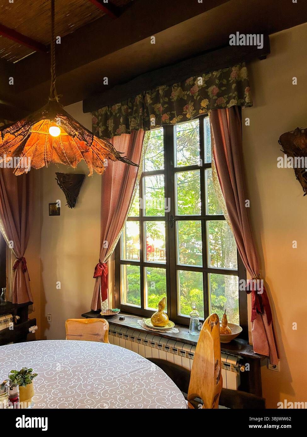 Warmer rustikaler Speisesaal mit Holzmöbeln, dekorativen Vorhängen und Tageslicht durch ein großes Fenster mit Blick auf den grünen Garten. - Smartphone-aufgenommenes Stockfoto