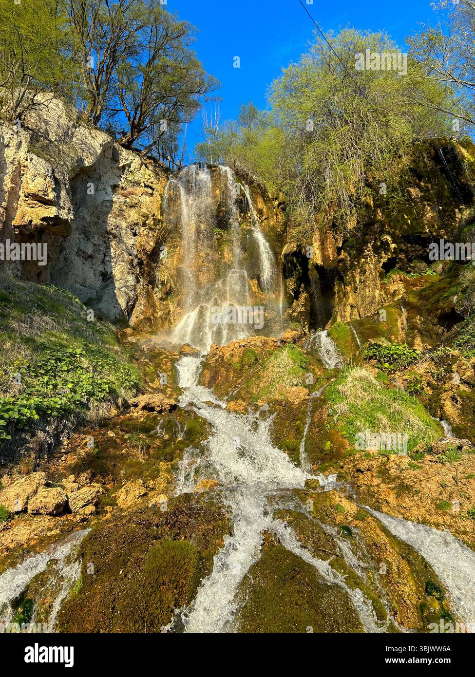 Atemberaubender Wasserfall, der unter einem hellblauen Himmel von moosigen Felsen umgeben ist von üppigem Grün und Bäumen. Ideal für Natur, Reisen und Outdoor - Smartphone-aufgenommenes Stockfoto