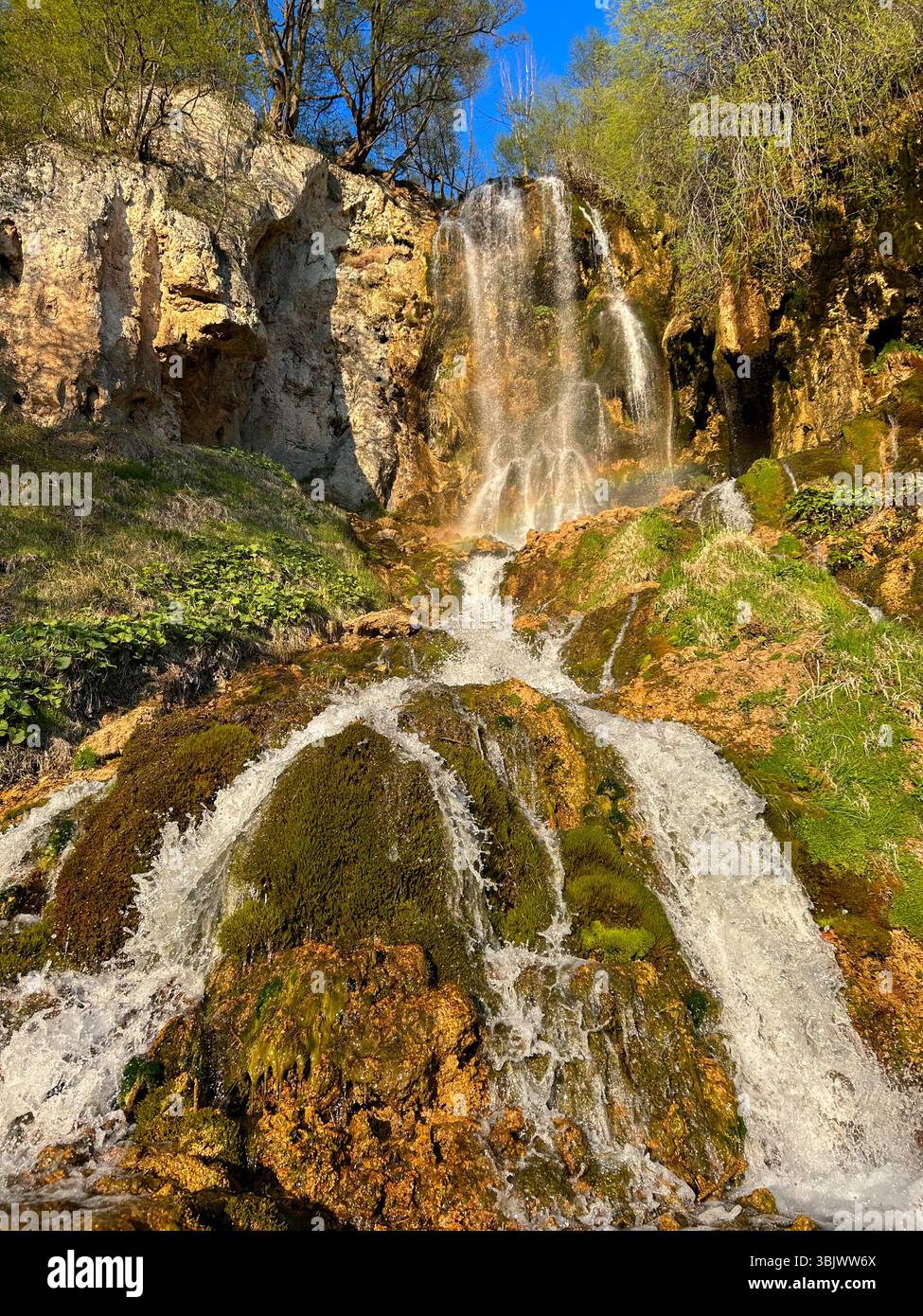 Wunderschöner natürlicher Wasserfall, der über moosbedeckte Felsen und Klippen fließt, umgeben von Grün unter einem klaren blauen Himmel. Perfekt für Natur, Reisen und - Smartphone-aufgenommenes Stockfoto