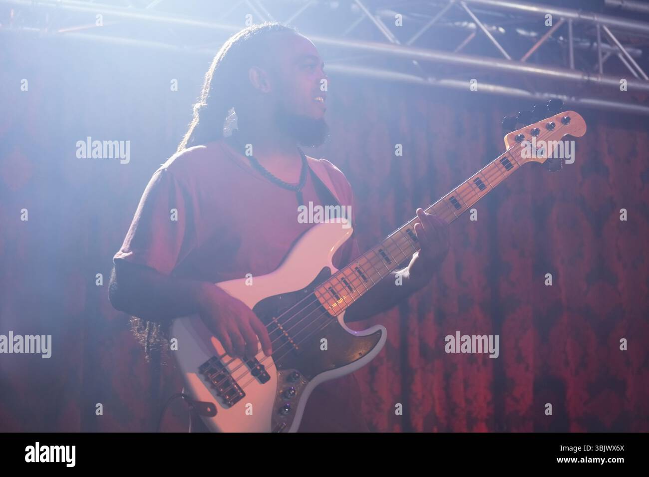 Musiker, der weiße E-Bass-Gitarre auf der Bühne unter Deckenleuchten spielt, roter gemusterter Vorhang Stockfoto