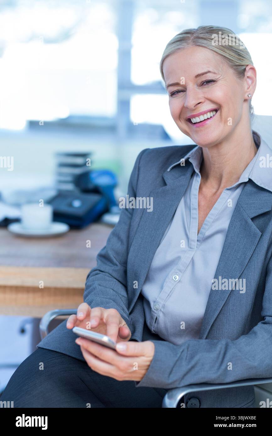 Geschäftsfrau mit Smartphone, lächelnd am Schreibtisch mit Telefon, Kaffeetasse, Akten, Milchfenster Stockfoto