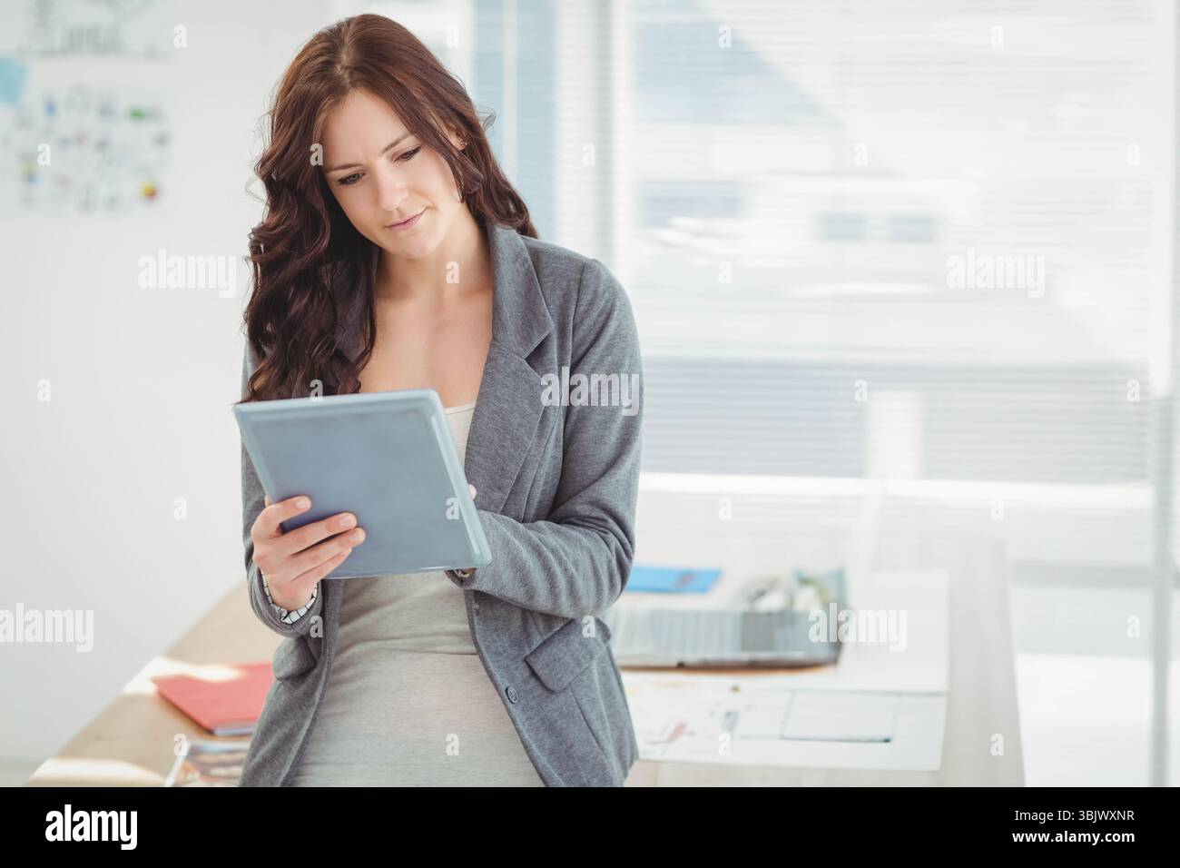 Tablet mit verstreuten Papieren auf dem Schreibtisch, Aktenablage unter horizontalen Jalousien in der Nähe der Pinnwand Stockfoto