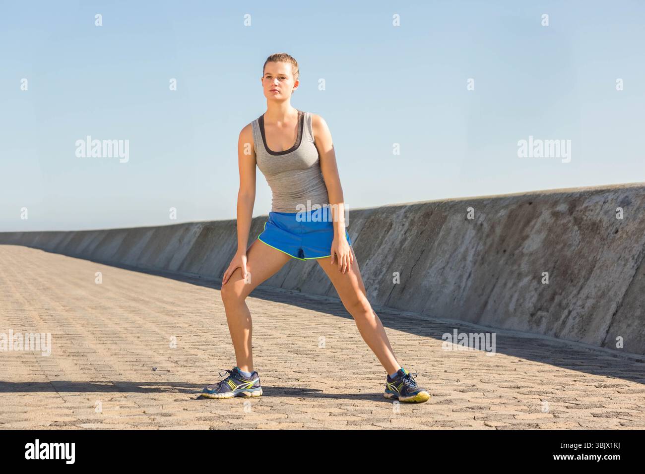 Frau, die seitlich Stretch auf der Promenade an der Uferwand trägt, trägt Tanktop, Shorts und Laufschuhe Stockfoto