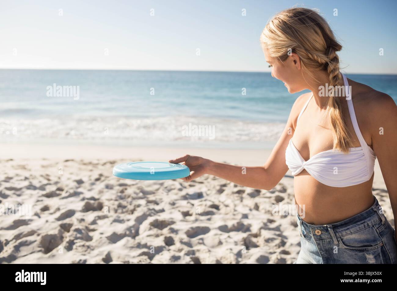 Erwachsene Frau, die am Sandstrand in der Nähe der Küste steht und eine hellblaue fliegende Scheibe unter klarem Himmel hält Stockfoto