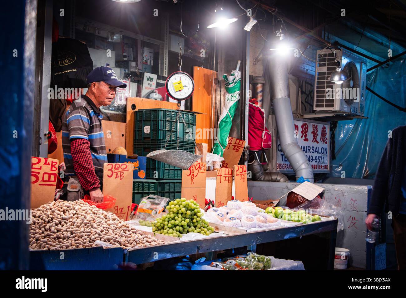 der asian Street Vendor am Night Time Market Stand verkauft Nüsse und Früchte, geschriebene Schilder und Hängewaage in der belebten urbanen Umgebung in Chinatown New York Stockfoto