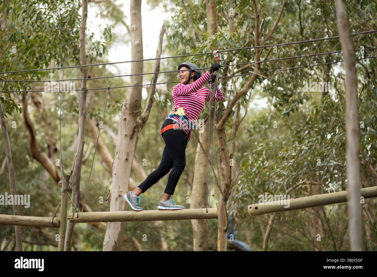 Afroamerikanerin, die ein Gurtzeug trägt, das auf einem Balken balanciert, das Oberkabel im Seilgarten hält Stockfoto