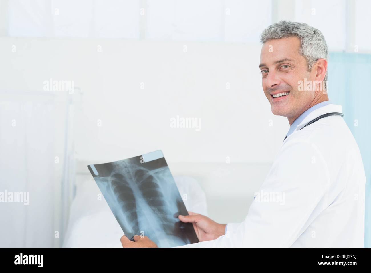 Arzt mittleren Alters mit Thoraxröntgenfilm mit Stethoskop in der Nähe des Patientenbettes im Krankenhauszimmer Stockfoto