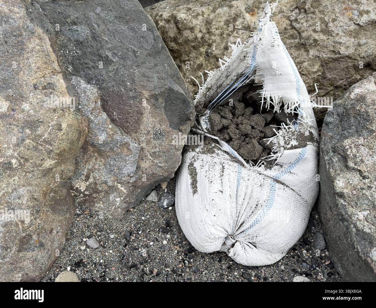 Abgerissene Sandsäcke gefüllt mit Kies und Sand, die zwischen Küstenblöcken platziert werden, um Erosionsschutz oder provisorische Absperrung zu gewährleisten. Keine Personen. Stockfoto
