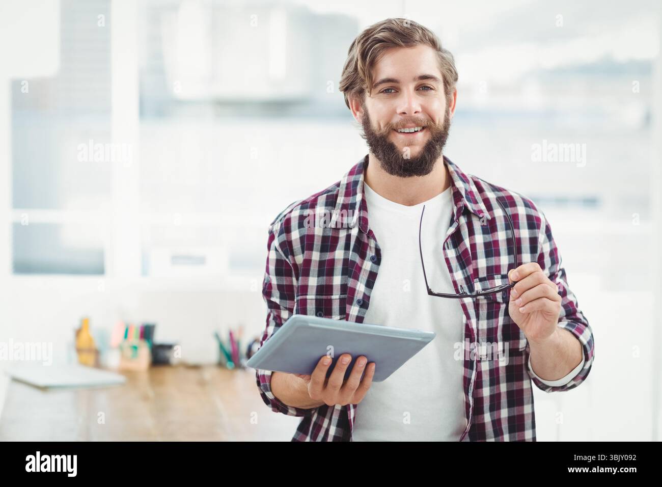 Bärtiger Mann mit Tablet und Brille in einem modernen Büro mit Holzschreibtisch und Stiften in der Nähe der Skyline Stockfoto