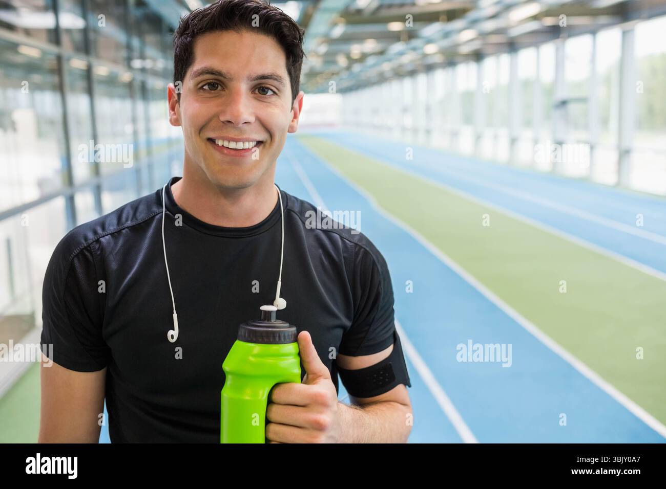 Männlicher Athlet in schwarzem Hemd, der auf der Schiene steht und eine grüne Wasserflasche mit Ohrhörern hält, Kopierraum Stockfoto