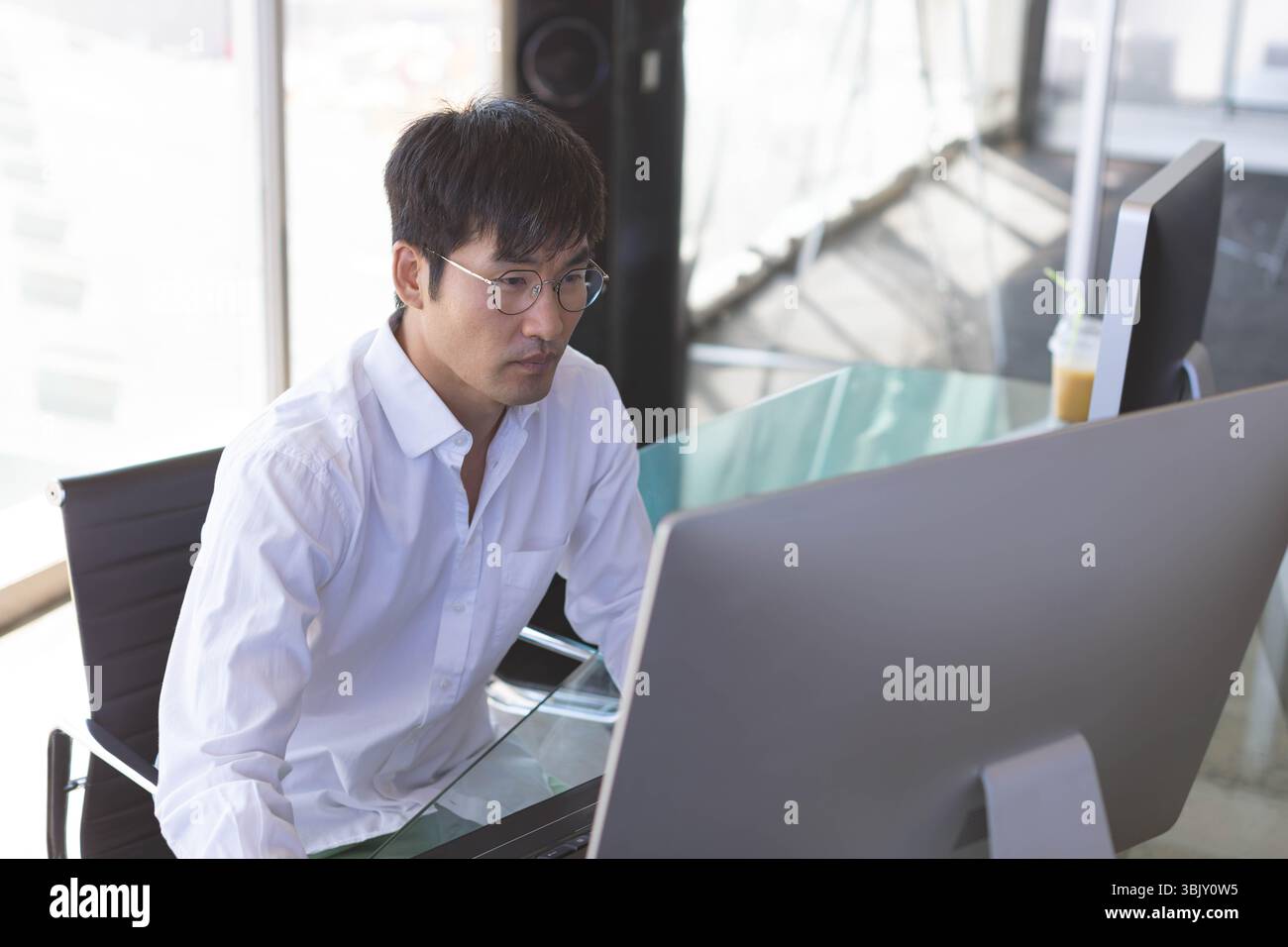 Computerbildschirm mit Bildschirm, Tasse und Lautsprecher, der auf dem Schreibtisch neben raumhohen Fenstern liegt Stockfoto