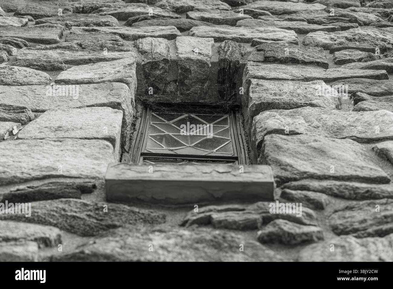Schwarzweißfoto eines kleinen Fensters in einer Steinmauer, von unten betrachtet, mit geometrischem Design und rauer gemauerter Textur, historischem Architekturdetail. Stockfoto Schwarzweißfoto eines kleinen Fensters in einer Steinmauer, von unten betrachtet, mit geometrischem Design und rauer gemauerter Textur, historischem Architekturdetail. Stockfoto