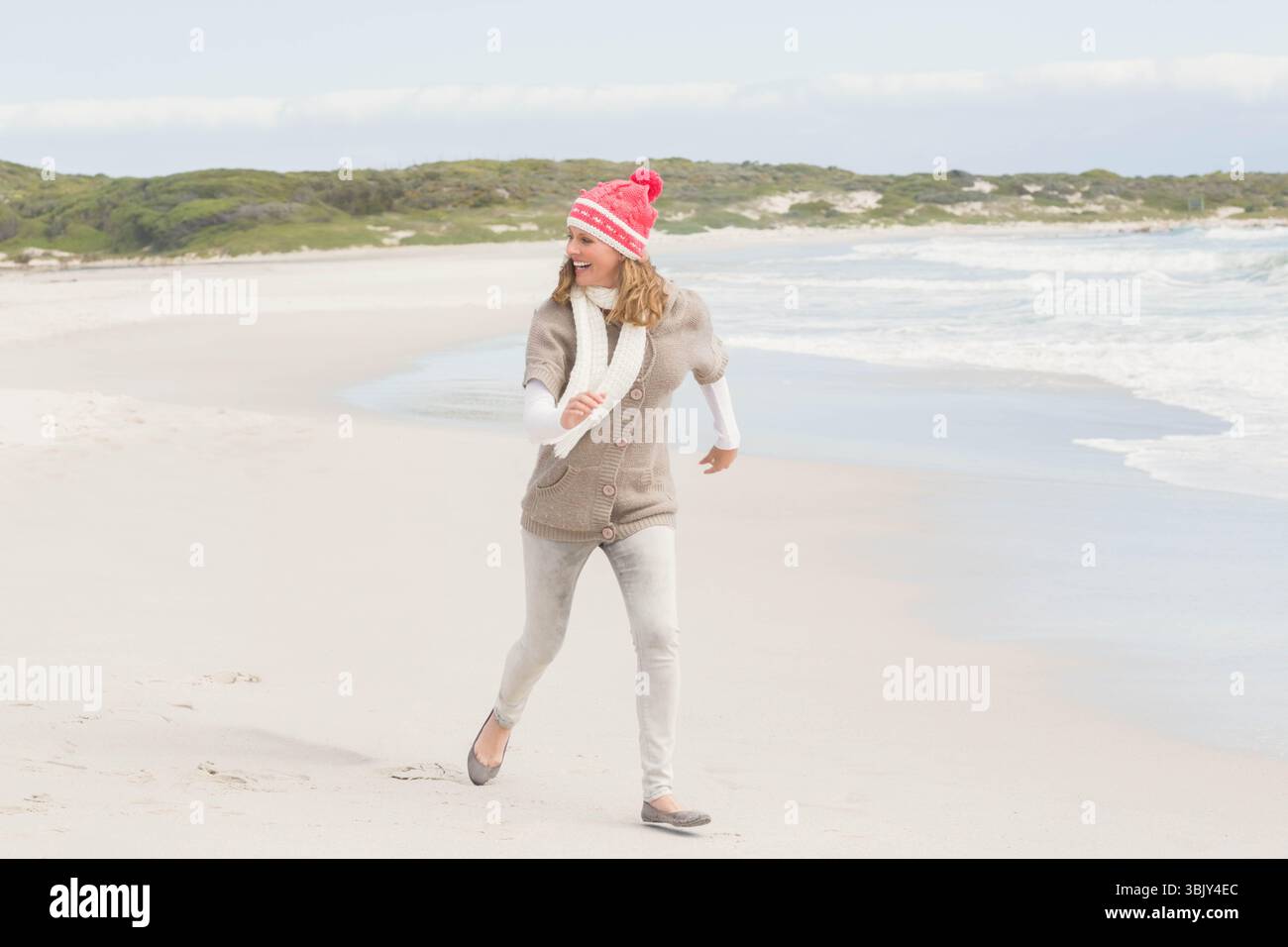 Frau, die am weißen Sandstrand läuft, trägt rote Strickmütze, weißen Schal, mit grüner Dünenlandschaft Stockfoto