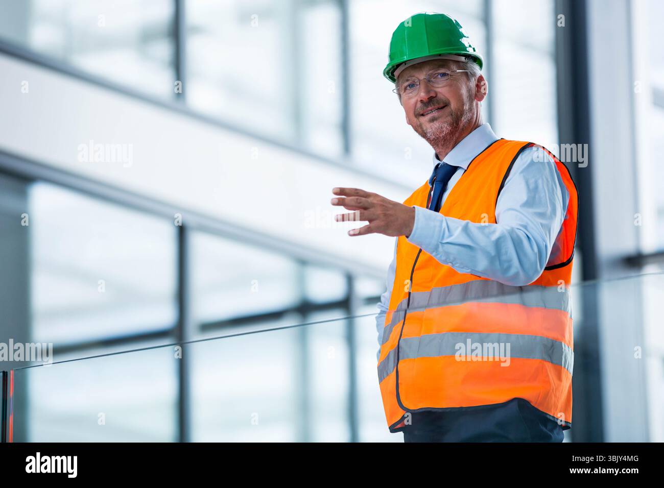 Der Manager trägt grünen Helm und orangefarbene Weste und bewegt sich hinter dem Glasgeländer im Atrium nach vorne Stockfoto