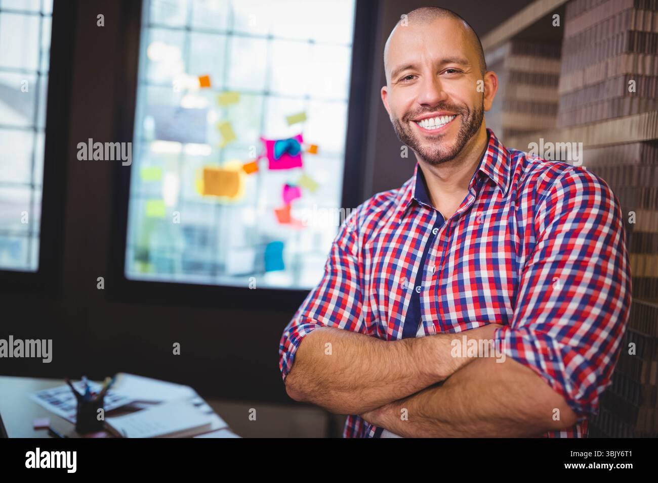 Büroangestellter mit überkreuzten Armen neben Schreibtischpapieren, bunte Haftnotizen am Fenster Stockfoto