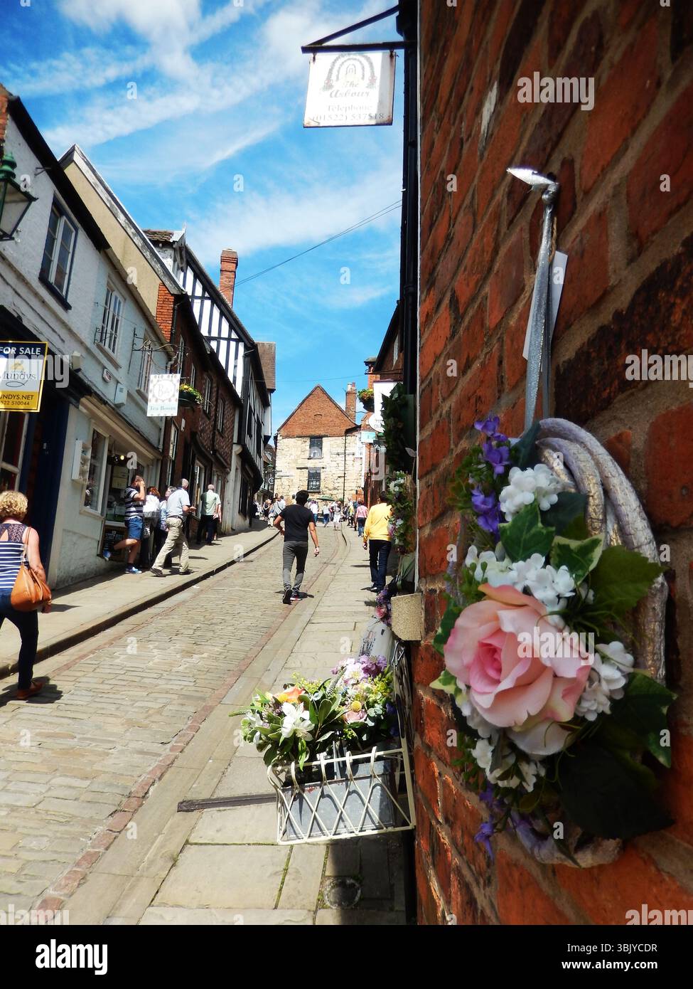 Historische Kopfsteinpflasterstraße gesäumt mit Geschäften und Blumendekorationen an einem sonnigen Tag. Lincoln, England Stockfoto