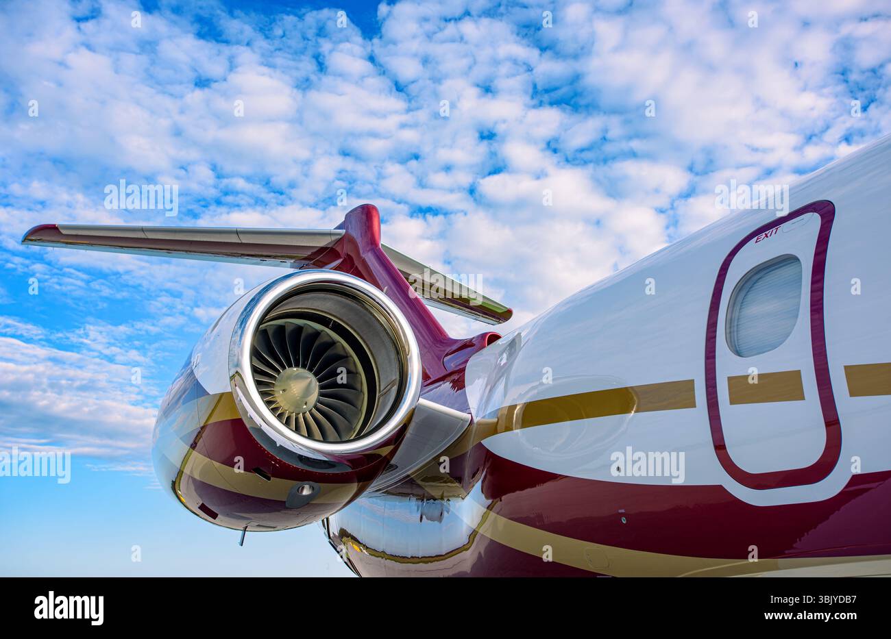 Seitenansicht des luxuriösen Business-Jets mit rot-goldener Lackierung. Jet-Motor, Rumpf und T-Tail sichtbar unter einem leuchtend blauen Himmel mit verstreuten Wolken. Stockfoto