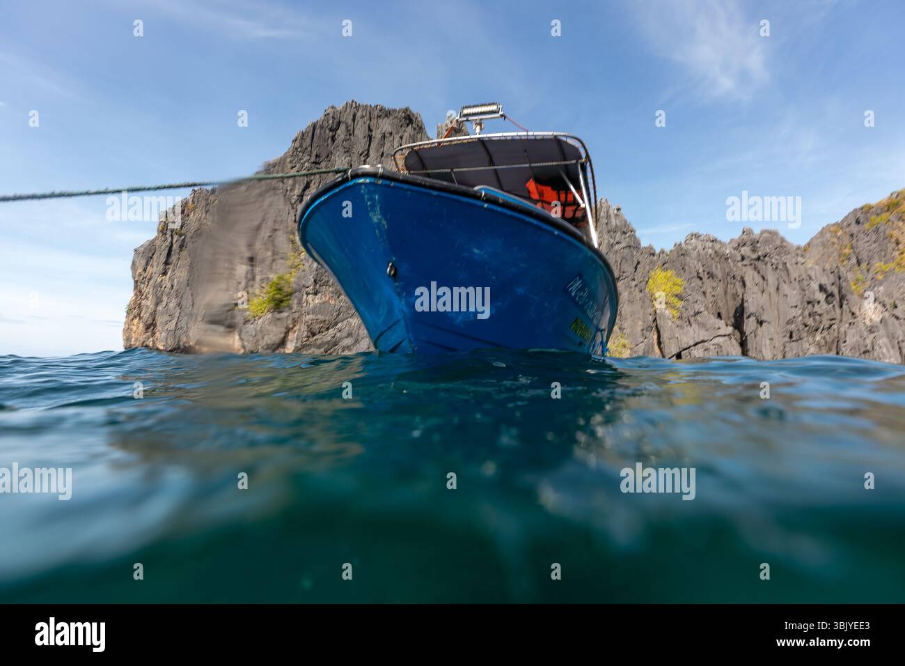 Ein blaues Boot liegt nahe einer felsigen Insel vor Anker, wobei die Kamera halb unter Wasser positioniert ist, um sowohl den Schiffsrumpf als auch das umliegende Meer zu erfassen. Stockfoto