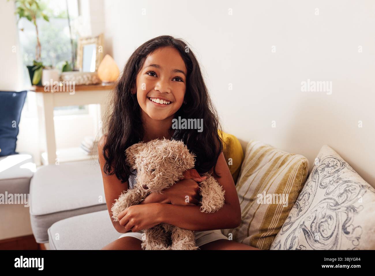 Porträt eines glücklichen Mädchens, das auf einem Sofa im Wohnzimmer sitzt und Teddybär umarmt Stockfoto