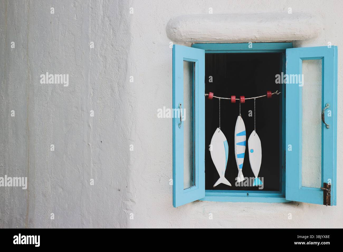 Pastellblaues Fenster mit hängendem Fischmobil an weißer Wand, mediterraner Stil mit Kopierraum auf der linken Seite. Minimales Wohndekor an der Küste. Stockfoto