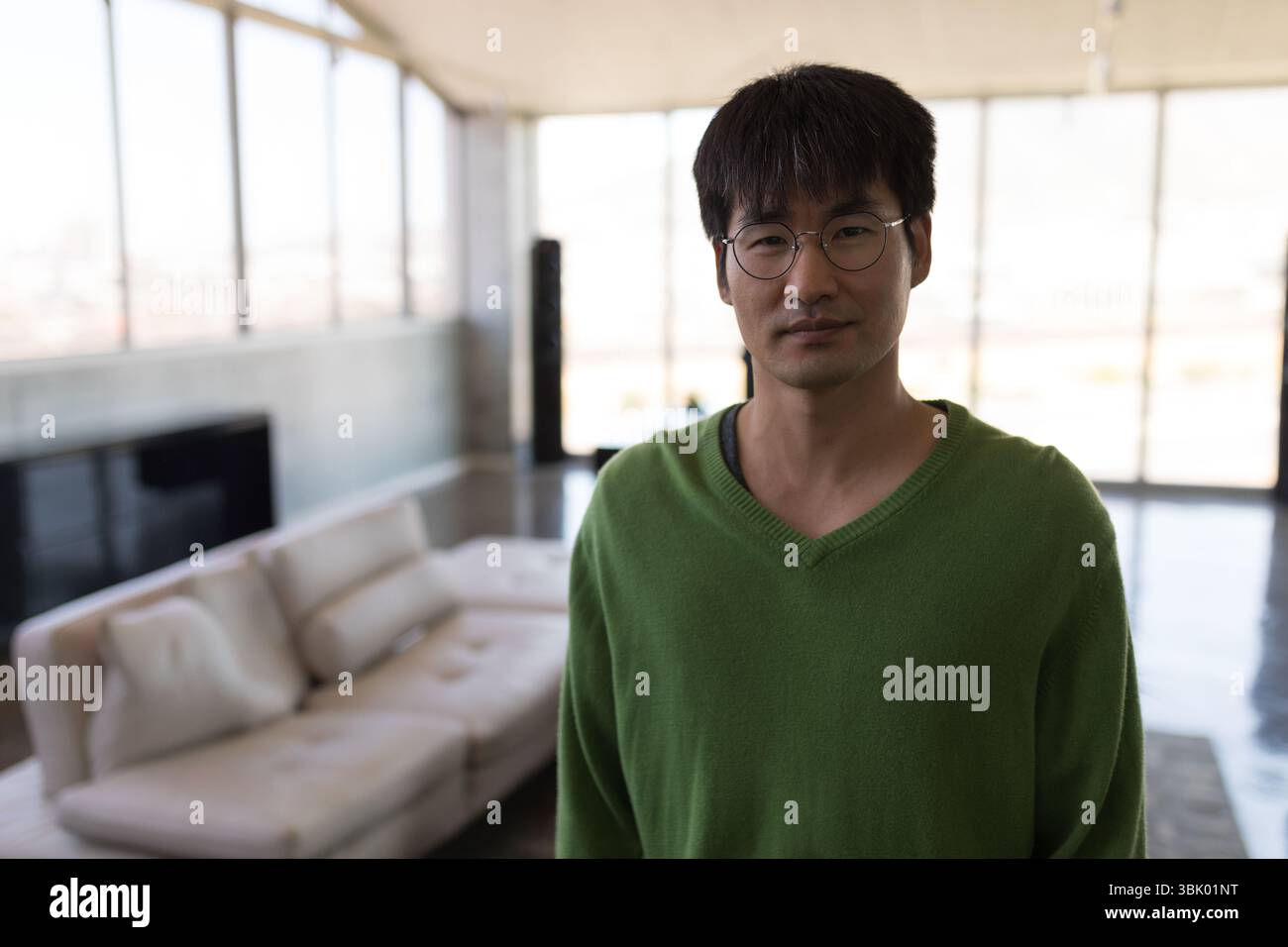 Asiatischer Mann mit grünem Pullover und Brille, der in der Lounge mit hohen Fenstern neben einem Ledersofa steht Stockfoto